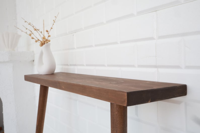 May include: A brown wooden console table with a rectangular top and four legs. A white vase with a unique shape holds dried branches with small, round, beige buds. The table is set against a white tiled wall.