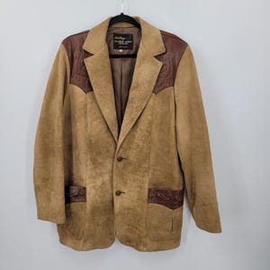 May include: A tan suede blazer with brown leather accents on the shoulders and pockets. The blazer has a notched lapel, two buttons, and a brown interior lining. The label reads "Joe Stage Leather Jacket".
