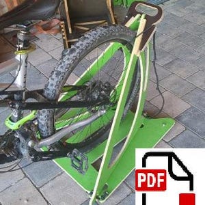 Op de afbeelding: Een groene houten fietsstandaard met een fietswiel vastgezet. De standaard heeft een rechthoekige basis en een gebogen frame. De fiets is zwart en groen. Een PDF-pictogram staat rechtsonder.