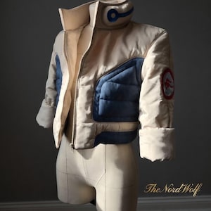 Könnte beinhalten: Cremefarbene, gesteppte Jacke mit blauen Akzenten und hohem Kragen. Die Jacke hat einen Reißverschluss, blaue Patches auf der Brust und einen runden Patch am Ärmel. Die Jacke wird auf einer Schneiderpuppe präsentiert.