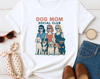 May include: White t-shirt featuring a graphic of three women with sunglasses and six dogs, with the text "DOG MOM SOCIAL CLUB" in red and blue. The shirt is styled with a pair of brown shoes, blue jeans, and a polka dot scarf.