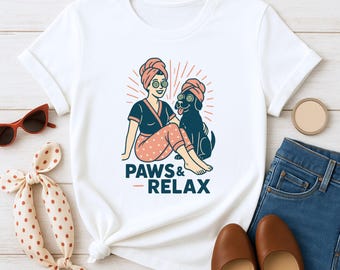 May include: White t-shirt featuring a cartoon illustration of a person and a dog relaxing with spa treatments. The graphic includes the text "PAWS & RELAX" in a blue and orange color scheme. The shirt is paired with jeans and brown shoes.