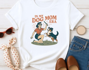 May include: White t-shirt featuring a retro illustration of a woman playing with a dog. The graphic includes the text "IN MY DOG MOM ERA" in orange and brown. The woman is wearing sunglasses, a red shirt, and a dark skirt.