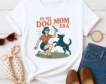 May include: White t-shirt featuring a retro illustration with the text "IN MY DOG MOM ERA." The graphic depicts a woman in sunglasses playing with a black dog. The shirt is styled with sunglasses, a scarf, jeans, and shoes.