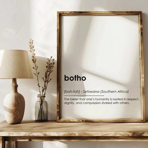 May include: A framed print with the word "botho" and its definition, displayed on a wooden table. A beige lamp and a small vase with dried plants are on the table. The print has a wooden frame.