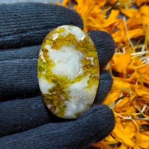 May include: An oval-shaped gemstone with a white base, featuring intricate patterns of yellow and green. The stone is held in a dark gray glove, with a background of orange flower petals.