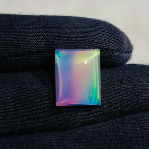 May include: A rectangular, iridescent gemstone with a polished surface. The gem displays a spectrum of colors, including pink, blue, green, and yellow, creating a shimmering effect. The stone is set against a dark background.