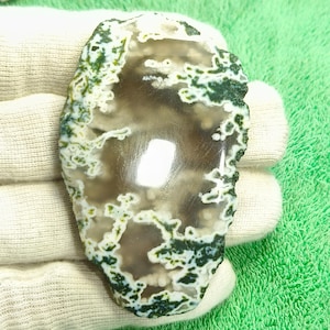 May include: A polished, oval-shaped gemstone with a unique pattern. The stone features a central area of light brown and gray, surrounded by white and green, creating a natural, organic design. The edges are rough, adding to its natural appeal.