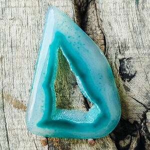Può includere: Una fetta di agata lucidata di forma triangolare color verde acqua, caratterizzata da un bordo esterno traslucido e una cavità geodica centrale rivestita di scintillanti formazioni cristalline chiare, con venature naturali visibili.