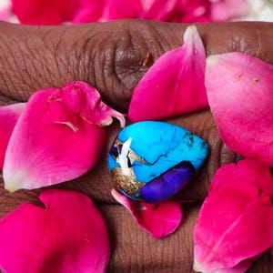 May include: A vibrant turquoise gemstone with gold accents and a deep purple base, surrounded by bright pink rose petals. The stone's unique color and texture are highlighted against the petals.