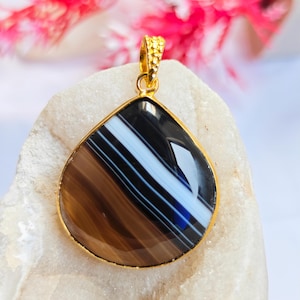 May include: A teardrop-shaped pendant featuring a polished banded agate stone with distinct layers of black, white, and brown. The gemstone is encased in a thin gold-toned metal bezel setting and includes a textured gold-toned metal bail at the top.