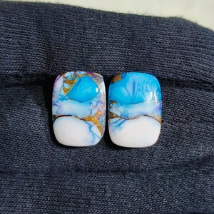 May include: Two rectangular cabochon gemstones with a striking design. The stones feature a combination of white, gold, and shades of blue, creating a unique and eye-catching appearance. The gemstones are likely for jewelry making.