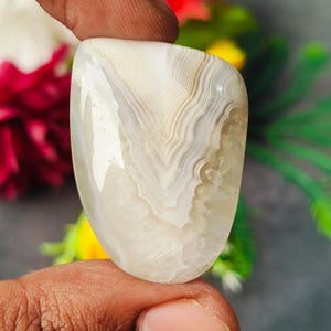 May include: A polished, shield-shaped agate gemstone featuring translucent white and cream-colored banding. The stone displays intricate, concentric wavy layers that converge toward the center, with a smooth, glossy surface texture.