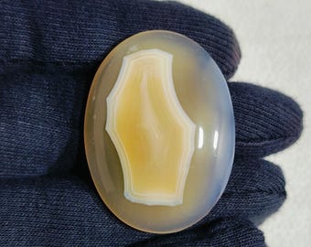 May include: Oval-shaped agate gemstone with a central, light-yellow design framed by white and a translucent, pale-blue outer layer. The polished stone is smooth and reflects light, showcasing natural patterns.