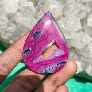 Può includere: Una gemma di agata lucida a forma di goccia con vivaci bande concentriche magenta e rosa e una cavità centrale di geode cristallino druzy. La pietra presenta bordi naturali traslucidi e inclusioni minerali viola scuro su tutta la superficie.