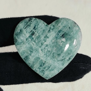 Puede incluir: Una piedra pulida en forma de corazón, de color verde mar translúcido. La piedra tiene una superficie texturizada con patrones cristalinos visibles. El corazón es liso y redondeado, con un ligero brillo.