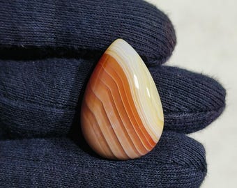 May include: A teardrop-shaped gemstone with bands of orange, brown, and white. The polished stone has a smooth, glossy surface. The gemstone is held in a dark blue fabric.