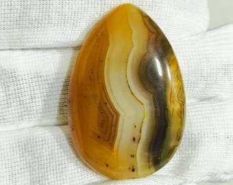 May include: A polished, teardrop-shaped gemstone with a mix of colors. The stone features bands of yellow, brown, white, and black, creating a layered effect. The surface is smooth and glossy, reflecting light. This gemstone is likely used for jewelry or decorative purposes.
