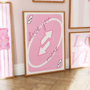 Puede incluir: Una l&aacute;mina decorativa rectangular en un marco de madera clara con un dise&ntilde;o de carta de juego rosa. El centro muestra dos flechas blancas que apuntan en direcciones opuestas, con el texto cursivo Love you more escrito en diagonal sobre el fondo rosa.
