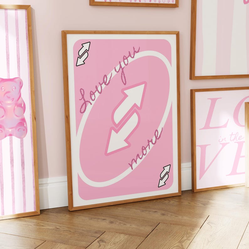 Puede incluir: Una l&aacute;mina decorativa rectangular en un marco de madera clara con un dise&ntilde;o de carta de juego rosa. El centro muestra dos flechas blancas que apuntan en direcciones opuestas, con el texto cursivo Love you more escrito en diagonal sobre el fondo rosa.