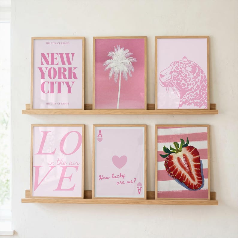 Puede incluir: Seis l&aacute;minas enmarcadas dispuestas en dos estantes de madera. La fila superior muestra un p&oacute;ster rosa de Nueva York, una palmera blanca sobre fondo rosa y un leopardo rosa. La fila inferior muestra un p&oacute;ster rosa de Love, un as de corazones y una fresa.