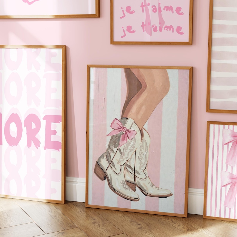 Puede incluir: Una l&aacute;mina de arte enmarcada que presenta una ilustraci&oacute;n pintada de una pierna inferior con una bota de estilo western blanca con un lazo rosa atado en el lateral. El fondo consiste en rayas verticales de color rosa claro y blanco, marco de madera.