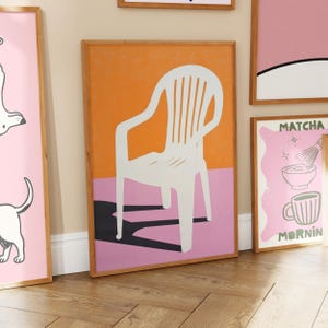 May include: A framed art print featuring a white chair against an orange and pink background. The chair's shadow is black. Other framed prints with pink backgrounds and illustrations are visible.