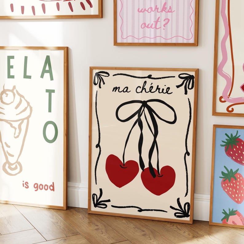 May include: A collection of framed art prints with a vintage aesthetic. One print features the text "ma ch&eacute;rie" above a pair of red heart-shaped cherries with black stems and a bow. Other prints include an ice cream cone, strawberries, and the phrase "works out?"