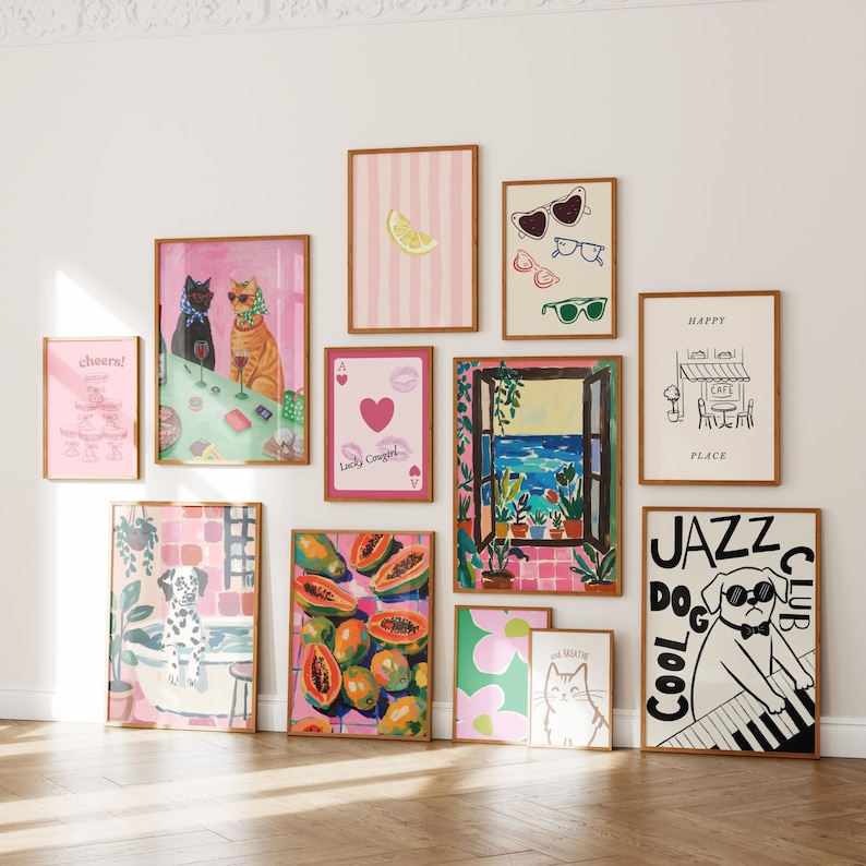 May include: A selection of framed art prints featuring diverse illustrations. The prints depict cats, a lemon, sunglasses, a cafe, a dog playing piano, and tropical fruit. The colour scheme is mainly pink, green, and white, with brown frames.