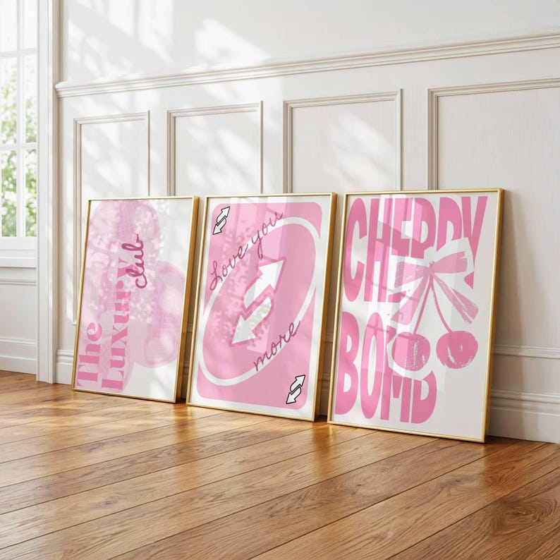 Puede incluir: Tres l&aacute;minas rectangulares en marcos dorados. La l&aacute;mina de la izquierda presenta formas abstractas rosas y el texto The Luxury club. La central parece una carta de juego rosa con flechas blancas y el texto Love you more. La de la derecha muestra cerezas rosas con un lazo y el texto CHERRY BOMB.