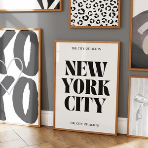 Pu&ograve; includere: Diverse stampe artistiche incorniciate con cornici in legno sono esposte su una parete grigia. Una stampa presenta il testo "NEW YORK CITY" in grassetto nero. Altre stampe hanno disegni astratti in bianco e nero.