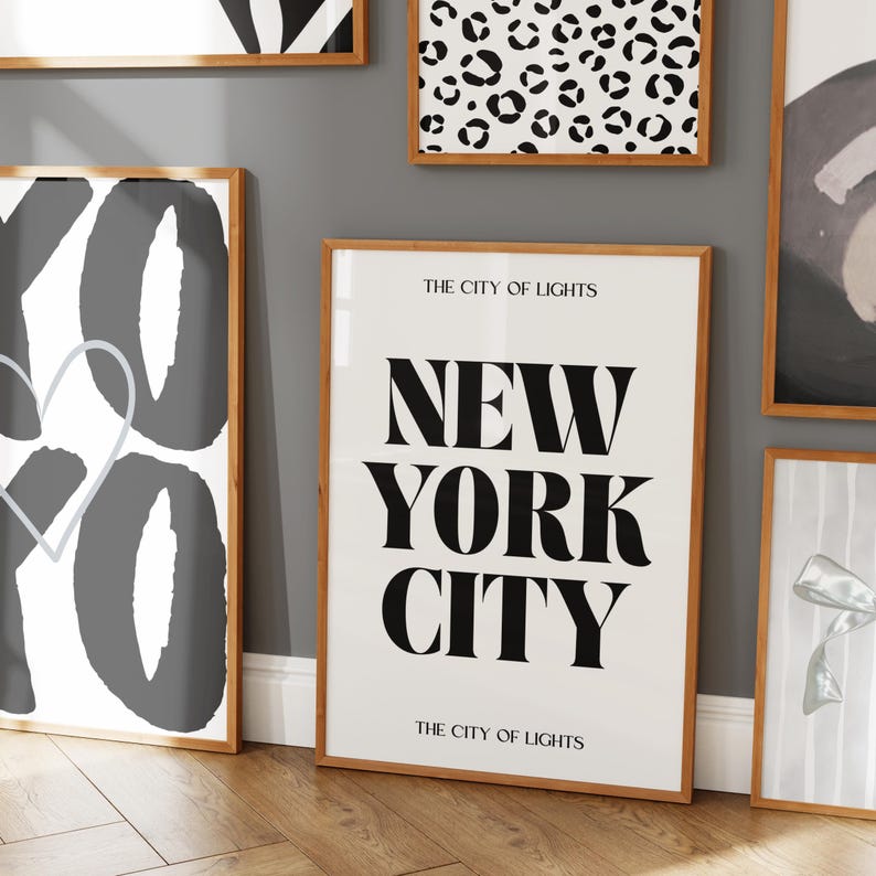 Pu&ograve; includere: Diverse stampe artistiche incorniciate con cornici in legno sono esposte su una parete grigia. Una stampa presenta il testo "NEW YORK CITY" in grassetto nero. Altre stampe hanno disegni astratti in bianco e nero.