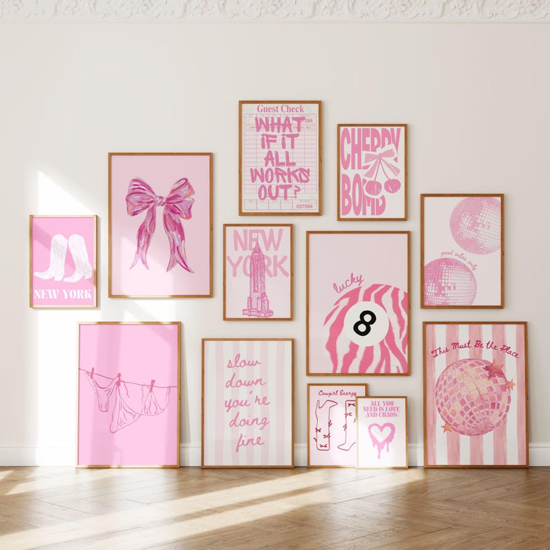May include: A gallery wall collection of eleven framed art prints featuring pink and white color schemes. Designs include a bow, New York skyline, disco balls, an eight ball, cowboy boots, laundry on a line, and various text-based graphics.