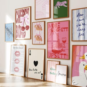 May include: A collection of framed art prints with various designs. Prints include text like "Cherry Bomb," "More Spaghetti," and "Slow Down." Other prints feature croissants, playing cards, and a leopard. The color palette is primarily pink, blue, and green.