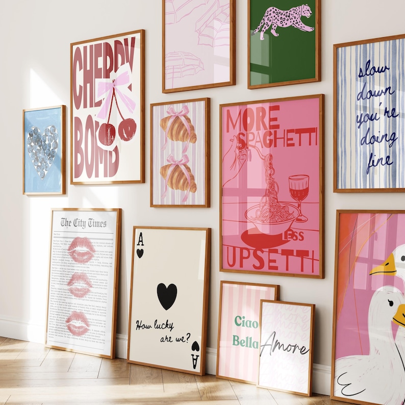 May include: A collection of framed art prints with various designs. Prints include text like "Cherry Bomb," "More Spaghetti," and "Slow Down." Other prints feature croissants, playing cards, and a leopard. The color palette is primarily pink, blue, and green.