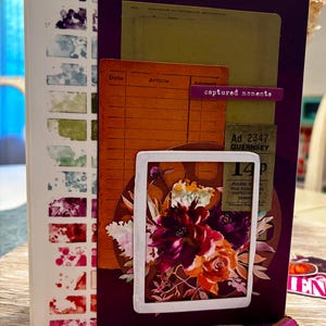 Peut inclure: Un journal avec une tranche blanche ornée d'une grille aquarelle colorée. La couverture violet foncé présente un collage superposé d'une fiche de registre orange vintage, d'un ticket vert et d'un cadre blanc entourant une illustration florale avec le texte captured moments.