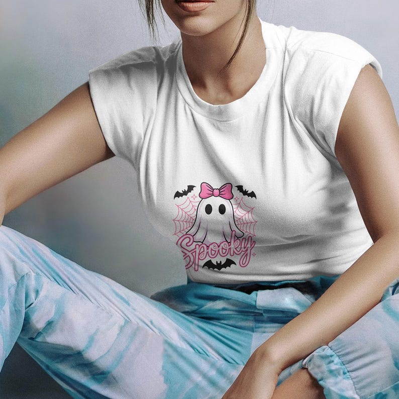 May include: White t-shirt featuring a cartoon ghost with a pink bow, surrounded by a spiderweb and bats. The word "Spooky" is written in pink script below the ghost. The shirt is part of a Halloween-themed collection.