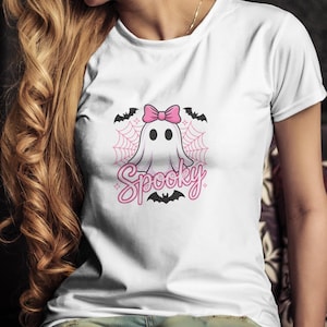May include: White t-shirt featuring a cartoon ghost with a pink bow, surrounded by a spiderweb and bats. The word "Spooky" is written in pink script below the ghost. A fun and festive design for Halloween.