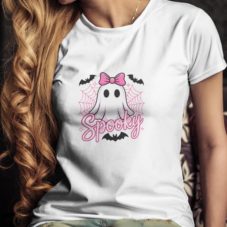 May include: White t-shirt featuring a cartoon ghost with a pink bow, surrounded by a spiderweb and bats. The word "Spooky" is written in pink script below the ghost. A fun and festive design for Halloween.