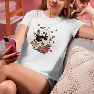 Puede incluir: Camiseta blanca con un dise&ntilde;o de Halloween. Un fantasma lee un libro con un gato negro, rodeado de flores, murci&eacute;lagos y hojas. El dise&ntilde;o tiene una est&eacute;tica vintage y retro.
