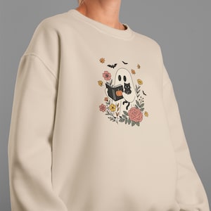 Puede incluir: Sudadera color crema con un dise&ntilde;o caprichoso de Halloween. Un fantasma amigable lee un libro con un gato negro, rodeado de flores, murci&eacute;lagos y calabazas. El dise&ntilde;o es en negro, naranja, rosa y amarillo.
