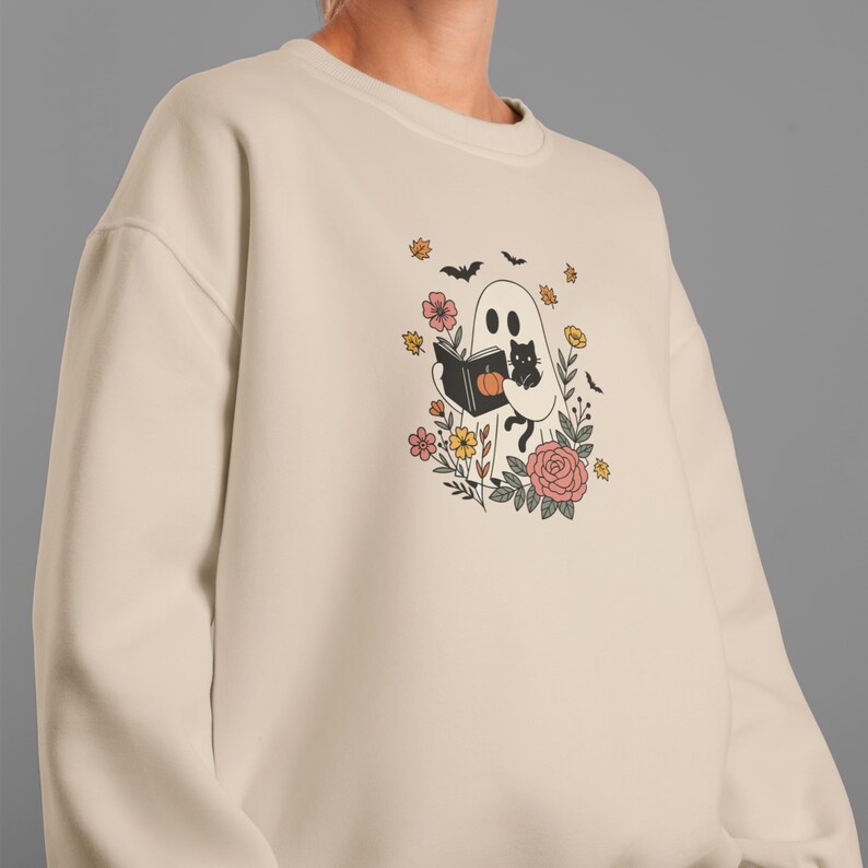 Puede incluir: Sudadera color crema con un dise&ntilde;o caprichoso de Halloween. Un fantasma amigable lee un libro con un gato negro, rodeado de flores, murci&eacute;lagos y calabazas. El dise&ntilde;o es en negro, naranja, rosa y amarillo.