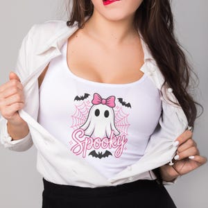 May include: White tank top with a cartoon ghost design. The ghost has a pink bow, and is surrounded by a pink spiderweb, bats, and the word "Spooky" in pink script. The tank top is part of a Halloween-themed collection.