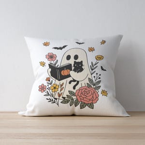 Puede incluir: Coj&iacute;n cuadrado blanco con un dise&ntilde;o de Halloween. Un fantasma lee un libro con una calabaza en la portada, sosteniendo un gato negro. El coj&iacute;n est&aacute; decorado con flores, hojas y murci&eacute;lagos.
