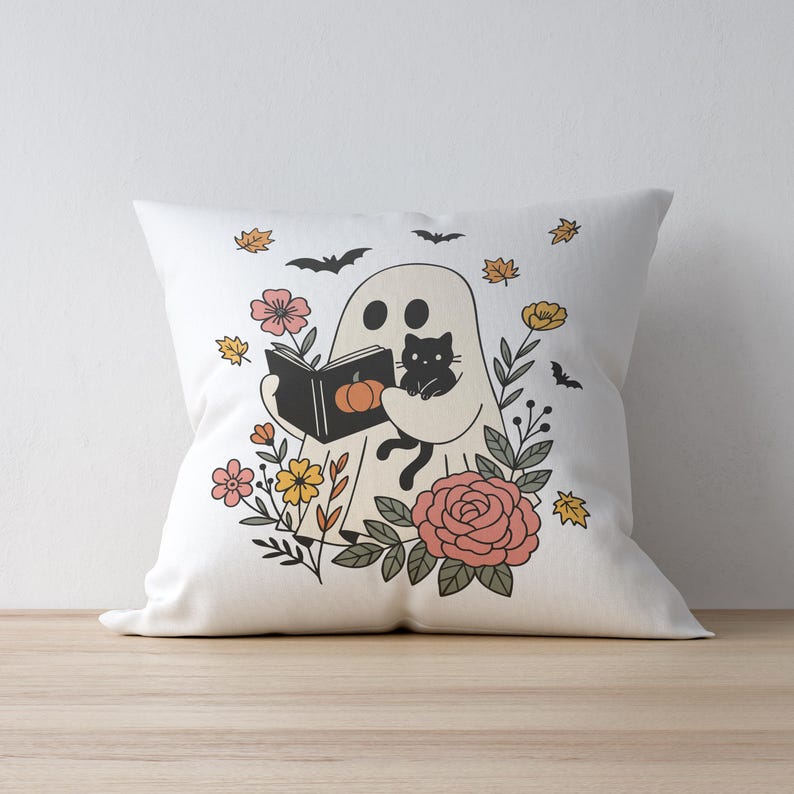 Puede incluir: Coj&iacute;n cuadrado blanco con un dise&ntilde;o de Halloween. Un fantasma lee un libro con una calabaza en la portada, sosteniendo un gato negro. El coj&iacute;n est&aacute; decorado con flores, hojas y murci&eacute;lagos.
