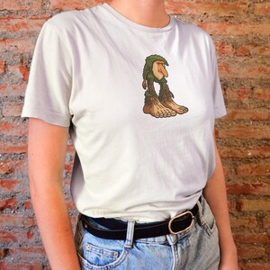 Op de afbeelding: Een lichtgrijs t-shirt met een cartoon troll ontwerp. De troll heeft een groene, bladerrijke kop, een lange neus en bruine benen en voeten. Het t-shirt heeft een eenvoudige ronde hals.