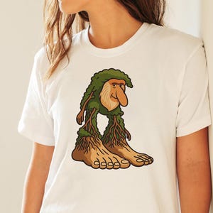 Op de afbeelding: Wit T-shirt met een cartoon troll illustratie. De troll heeft een groene, bladerrijke kop, een grote neus en boomachtige benen en voeten. Het ontwerp gebruikt groen, bruin en beige.