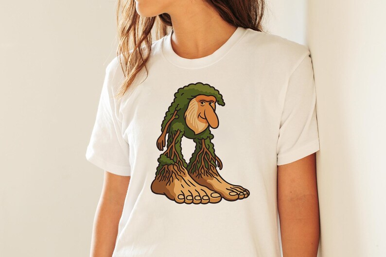 Op de afbeelding: Wit T-shirt met een cartoon troll illustratie. De troll heeft een groene, bladerrijke kop, een grote neus en boomachtige benen en voeten. Het ontwerp gebruikt groen, bruin en beige.