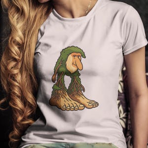 Op de afbeelding: Wit T-shirt met een cartoon illustratie van een personage met een groene, bladachtige kop en lichaam, en grote, wortelachtige voeten. Het personage heeft een lange neus en een vriendelijke uitdrukking. Het ontwerp is gecentreerd op het shirt.