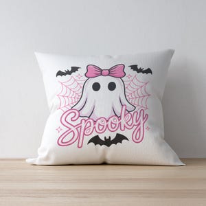 May include: White square pillow with a cartoon ghost wearing a pink bow. The word "Spooky" is written in pink script below the ghost, with bats and a spiderweb design. A Halloween-themed decorative pillow.